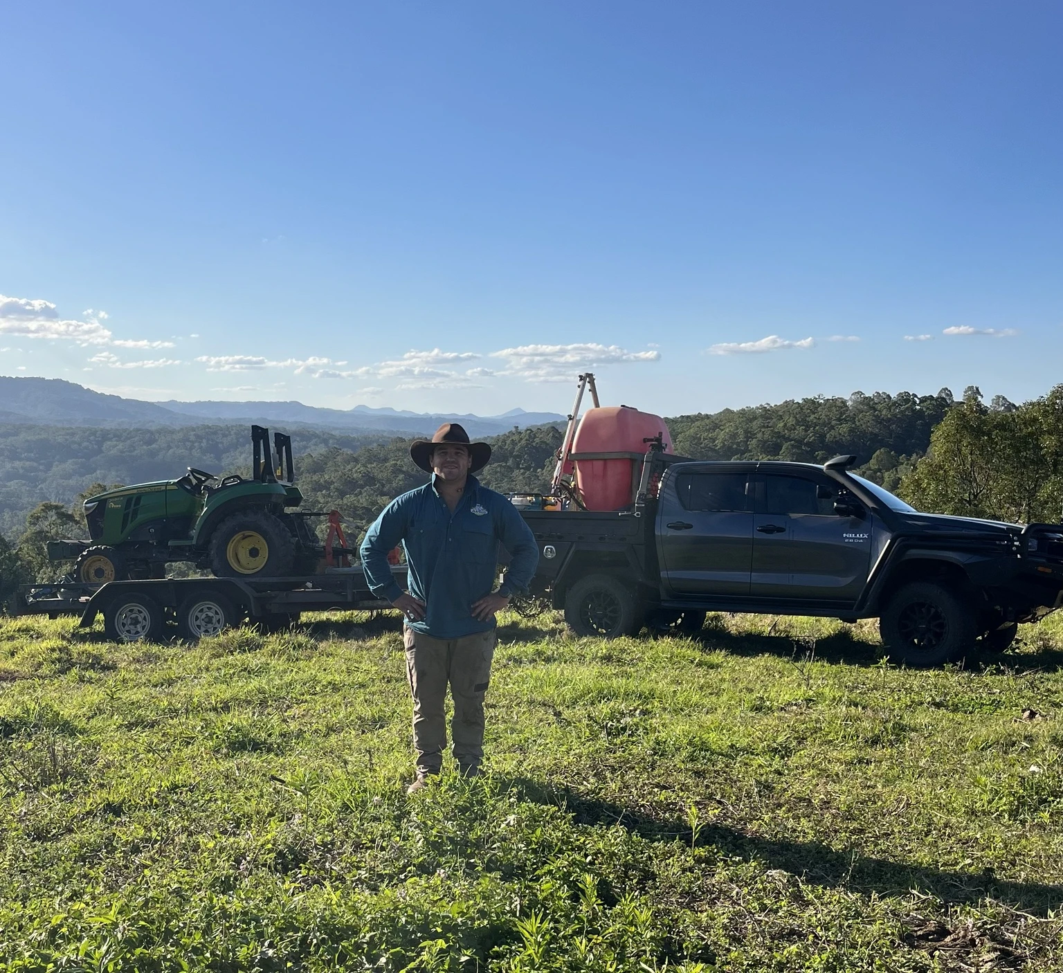 James standing in a paddock in front of his ute, John Deere tractor and spraying equipment, with hinterland views